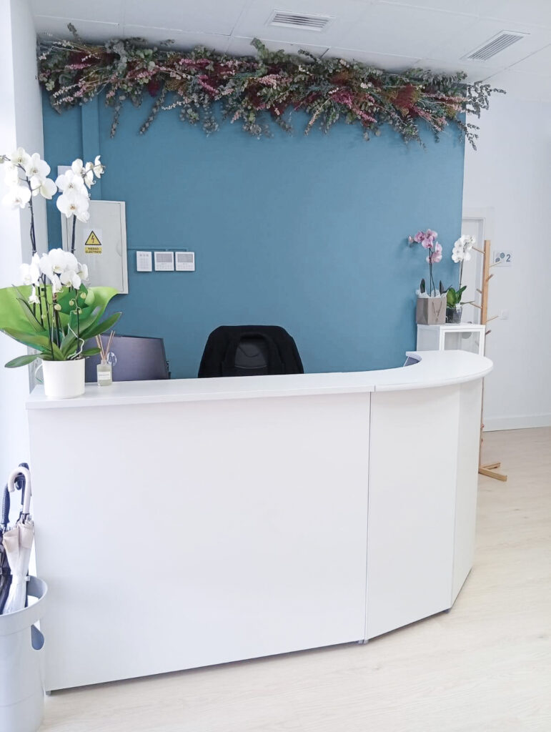 Recepción de la Clínica Thodos Granada con mostrador blanco curvo, pared azul decorada con flores y plantas, mostrando las instalaciones de la clínica