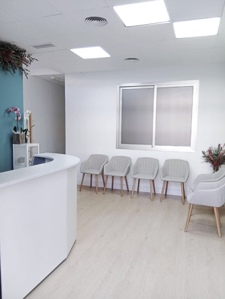 Recepción de la Clínica Thodos Granada con mostrador blanco curvo, iluminación moderna y zona de espera con sillas, mostrando las instalaciones de la clínica