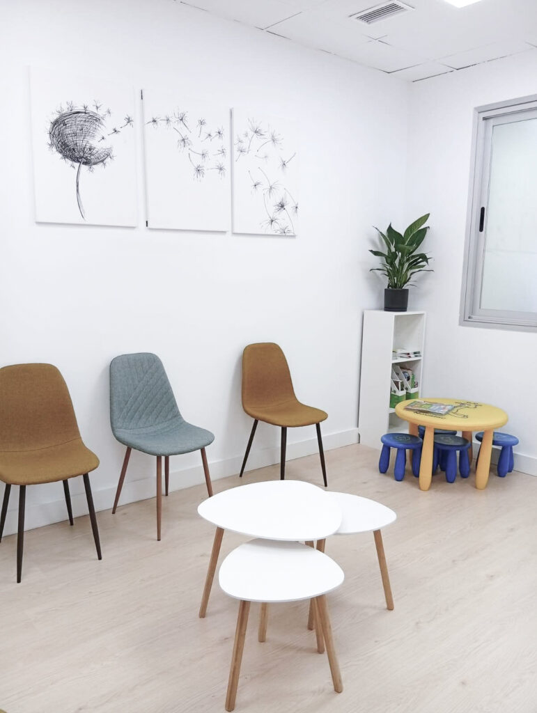 Sala de espera de la Clínica Thodos Granada con decoración de diente de león en la pared, sillas de colores y zona infantil con mesa pequeña, mostrando las instalaciones de espera de la clínica