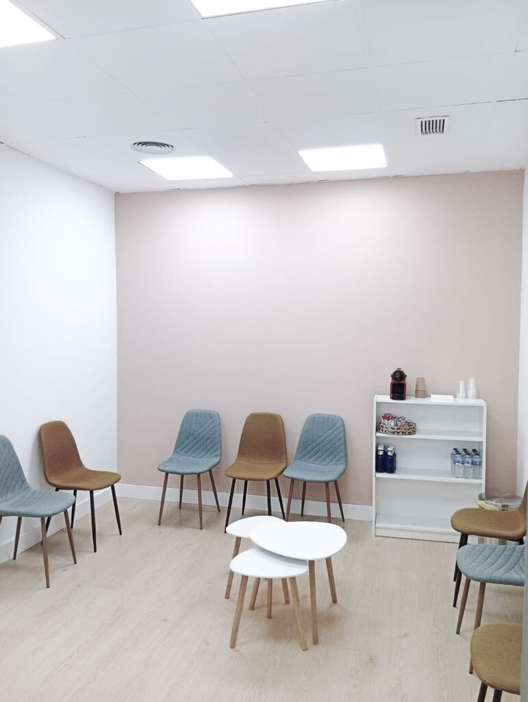 Sala de espera de la Clínica Thodos Granada con sillas de colores, pared de tono pastel y estantería con objetos decorativos, mostrando las instalaciones de espera de la clínica